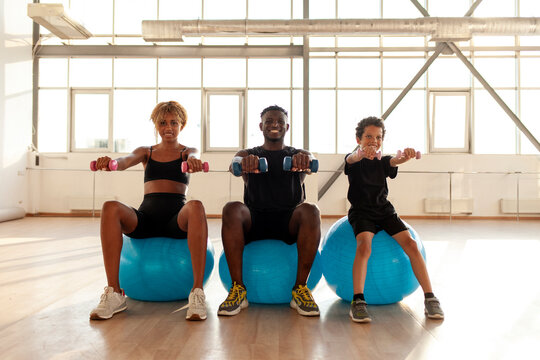 sporty african american family is doing pilates on fitness ball in the gym, boy with his parents is holding dumbbells - Powered by Adobe