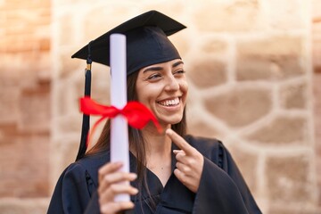 Young hispanic woman wearing graduated uniform pointing with finger to diploma at university