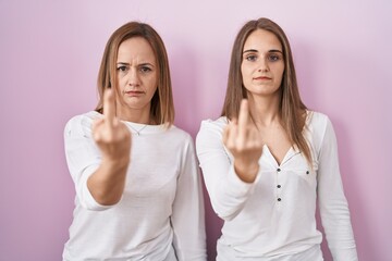 Middle age mother and young daughter standing over pink background showing middle finger, impolite and rude fuck off expression