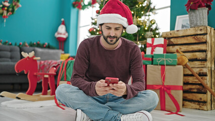 Obraz na płótnie Canvas Young hispanic man using smartphone celebrating christmas at home