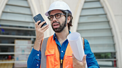 Young hispanic man architect holding blueprints sending voice message by smartphone at construction place