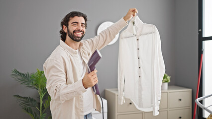 Young hispanic man ironing shirt with vertical iron machine smiling at laundry room