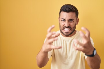 Fototapeta premium Handsome hispanic man standing over yellow background shouting frustrated with rage, hands trying to strangle, yelling mad