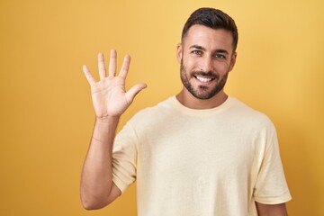 Handsome hispanic man standing over yellow background showing and pointing up with fingers number five while smiling confident and happy.