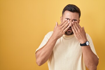 Handsome hispanic man standing over yellow background rubbing eyes for fatigue and headache, sleepy...