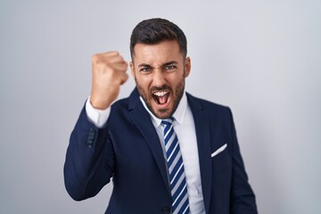 Handsome hispanic man wearing suit and tie angry and mad raising fist frustrated and furious while shouting with anger. rage and aggressive concept.