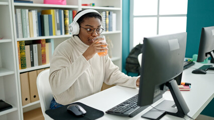 African american woman student using computer and headphones drinking orange juice at library university