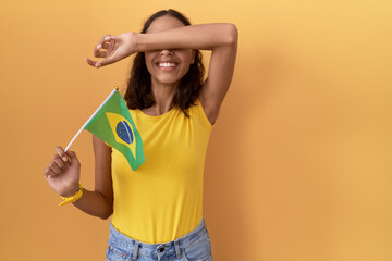 Young hispanic woman holding brazil flag smiling cheerful playing peek a boo with hands showing face. surprised and exited