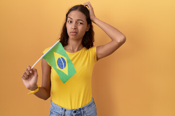 Young hispanic woman holding brazil flag confuse and wondering about question. uncertain with doubt, thinking with hand on head. pensive concept.
