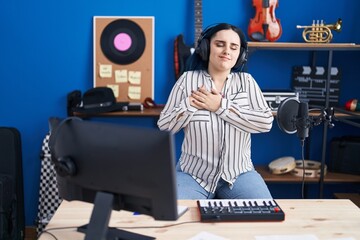Fototapeta na wymiar Young modern girl with blue hair at music studio wearing headphones smiling with hands on chest, eyes closed with grateful gesture on face. health concept.