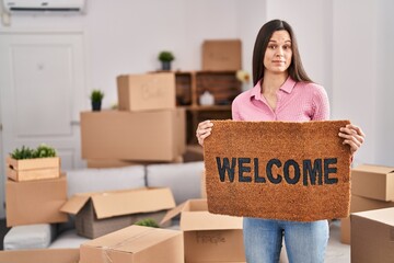 Young hispanic woman holding welcome doormat at new home smiling looking to the side and staring...