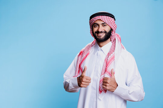 Arab man wearing islamic clothes giving thumbs up and showcasing cheerful expression portrait. Smiling muslim person dressed in traditional robe and headscarf and looking at camera