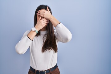 Young brunette woman standing over blue background covering eyes and mouth with hands, surprised...