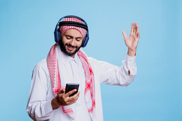 Muslim man wearing ghutra headscarf and wireless headphones changing song in playlist on smartphone. Arab listening to music in earphones, gesticulating and using mobile phone