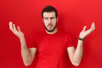 Young hispanic man wearing casual red t shirt clueless and confused expression with arms and hands raised. doubt concept.