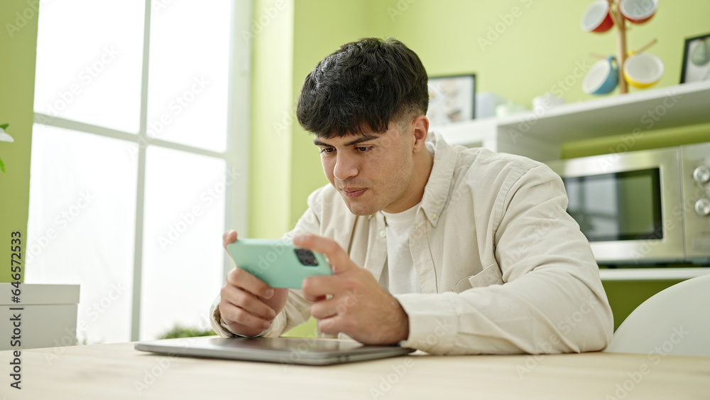 Sticker Young hispanic man playing video game sitting on table at dinning room