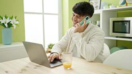 Young hispanic man using laptop talking on smartphone sitting on table at dinning room