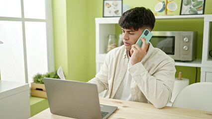Young hispanic man using laptop talking on smartphone at dinning room