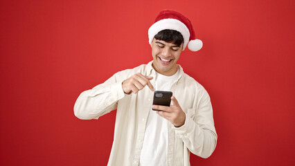 Young hispanic man wearing christmas hat pointing to smartphone over isolated red background