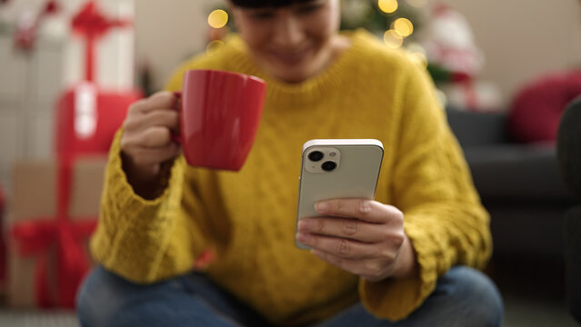 Young chinese woman using smartphone drinking coffee by christmas tree at home