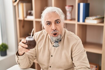 Middle age man with grey hair drinking mate infusion scared and amazed with open mouth for...