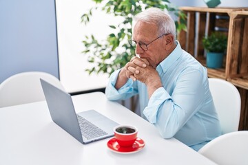 Senior grey-haired man using laptop and drinking coffee sitting on table at home