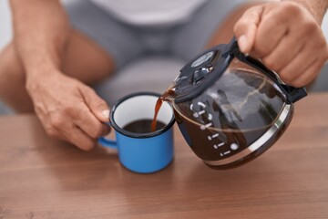 Middle age grey-haired man pouring coffee on cup sitting on sofa at home
