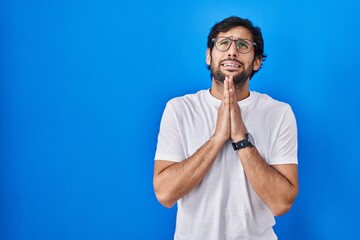 Handsome latin man standing over blue background begging and praying with hands together with hope expression on face very emotional and worried. begging.