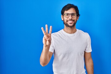 Fototapeta premium Handsome latin man standing over blue background showing and pointing up with fingers number three while smiling confident and happy.