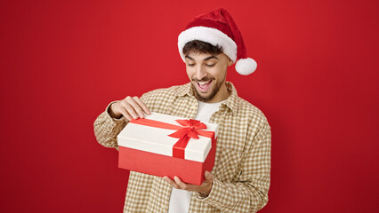 Young arab man wearing christmas hat unpacking gift over isolated red background