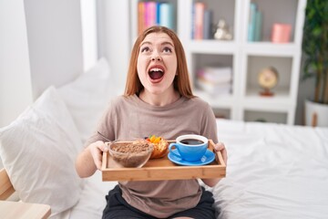 Redhead woman wearing pajama holding breakfast tray angry and mad screaming frustrated and furious, shouting with anger looking up.