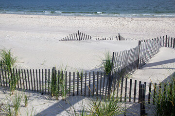 A wooden slated sand fence is partly buried in an ocean beach dune. There is sea grass growing...