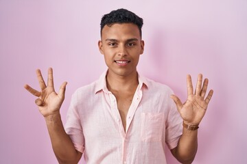 Young hispanic man standing over pink background showing and pointing up with fingers number nine...