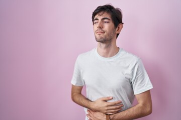 Young hispanic man standing over pink background with hand on stomach because indigestion, painful illness feeling unwell. ache concept.