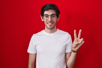 Naklejka premium Young hispanic man standing over red background showing and pointing up with fingers number two while smiling confident and happy.