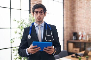Young hispanic man wearing doctor stethoscope at the office clueless and confused expression. doubt...
