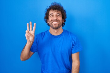 Naklejka premium Hispanic young man standing over blue background showing and pointing up with fingers number three while smiling confident and happy.