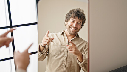 Young hispanic man looking on mirror pointing to fingers at home