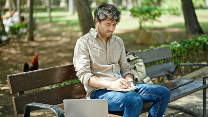 Young hispanic man writing on notebook using laptop at park