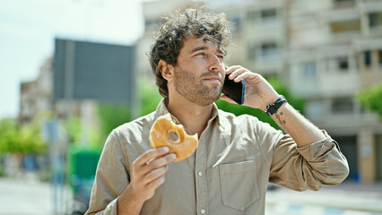 Young hispanic man eating doughnut talking on smartphone at street