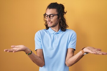 Young hispanic man standing over yellow background smiling showing both hands open palms, presenting and advertising comparison and balance