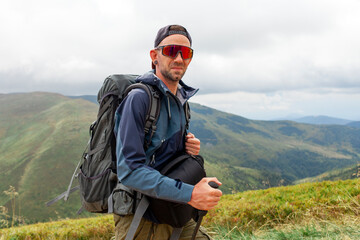 young man in orange jacket and glasses stands on the top of mountain with backpack and trekking...