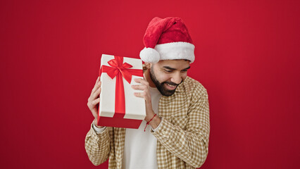 Young hispanic man smiling confident wearing christmas hat hearing gift sound over isolated red background