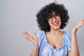 Young brunette woman with curly hair wearing glasses over isolated background celebrating crazy and amazed for success with arms raised and open eyes screaming excited. winner concept