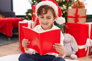 Fototapeta na wymiar Adorable hispanic boy reading book sitting on floor by christmas tree at home