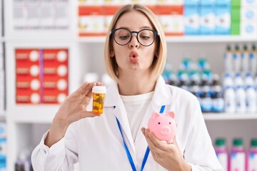 Young caucasian woman working at pharmacy drugstore holding pills an piggy bank making fish face with mouth and squinting eyes, crazy and comical.