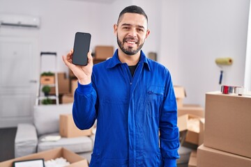 Hispanic man working on moving service showing smartphone screen looking positive and happy standing and smiling with a confident smile showing teeth