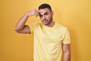 Fototapeta na wymiar Young hispanic man standing over yellow background pointing unhappy to pimple on forehead, ugly infection of blackhead. acne and skin problem