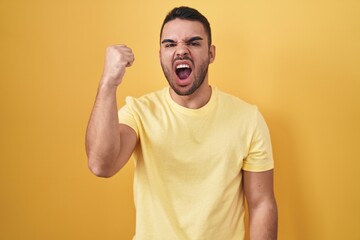 Fototapeta na wymiar Young hispanic man standing over yellow background angry and mad raising fist frustrated and furious while shouting with anger. rage and aggressive concept.