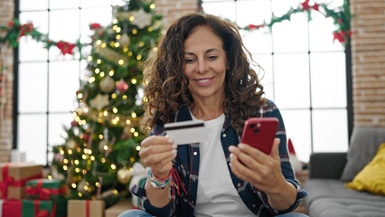 Middle age hispanic woman shopping with smartphone and credit card celebrating christmas at home